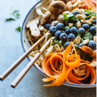 a bowl with chopsticks inside of thai noodles with blueberries, shredded carrots, peanuts, and mushrooms
