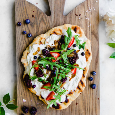 a flatbread with burrata cheese spread on top with berries and arugula on a wooden board