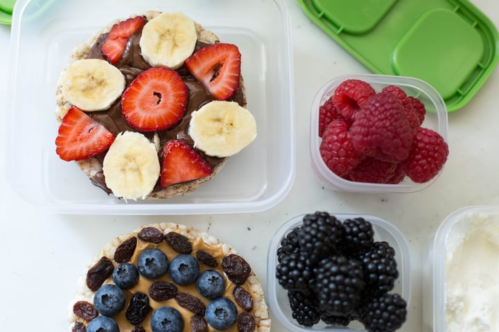 rice cake topped with blueberries, peanut butter, and raisins. and a rice cake topped with nutella, strawberries, and bananas with a side of raspberries and blackberries 