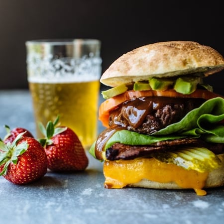 a hamburger with beef, bacon, avocado, tomatoes, and lettuce with strawberries on the side