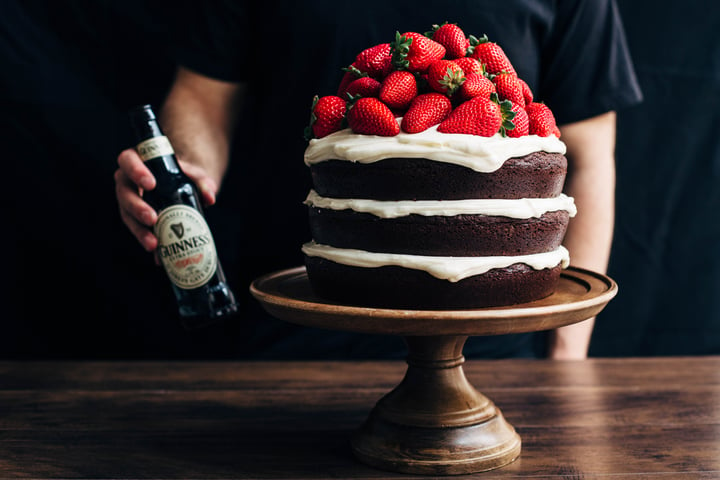 someone holding a Guinness beer next to a chocolate cake topped with strawberries