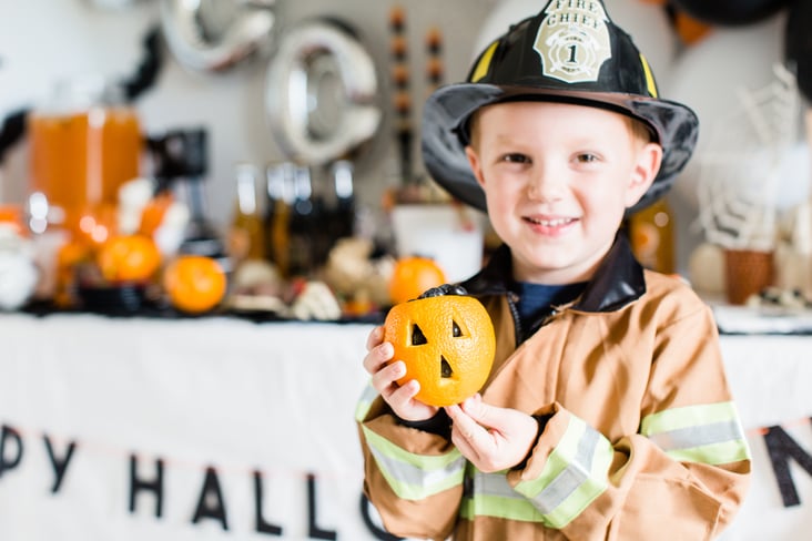 a child dressed as a firefighter holding an orange made to look like a jack-o-lantern