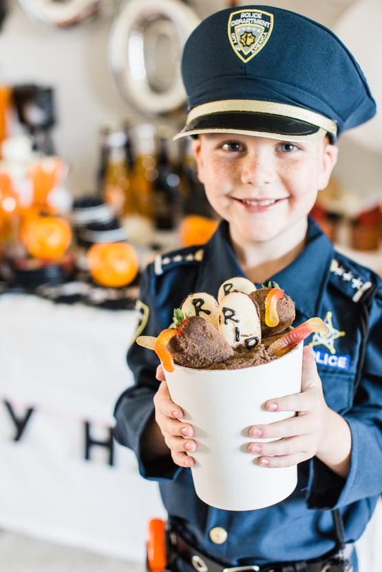 a child dressed as a police officer holding a Halloween dessert with gummy worms made to look like they're coming out of dirt 