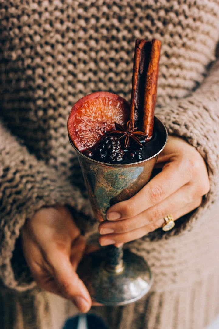 someone holding a metal wine cup filled with wine and garnished with blackberries, a cinnamon stick, and a slice of an orange