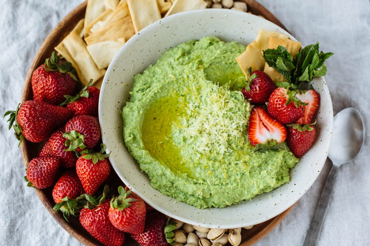 sweet pea hummus with a strawberry garnish and berries, crackers, and pistachios on the side