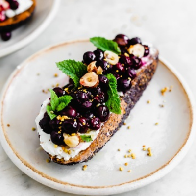 a slice of bread with goat cheese spread on topped with blueberries, hazelnuts, and mint leaves on a plate.
